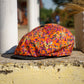 A flat cap with a vibrant orange, pink, and purple camouflage pattern, resting on a weathered stone ledge outdoors.