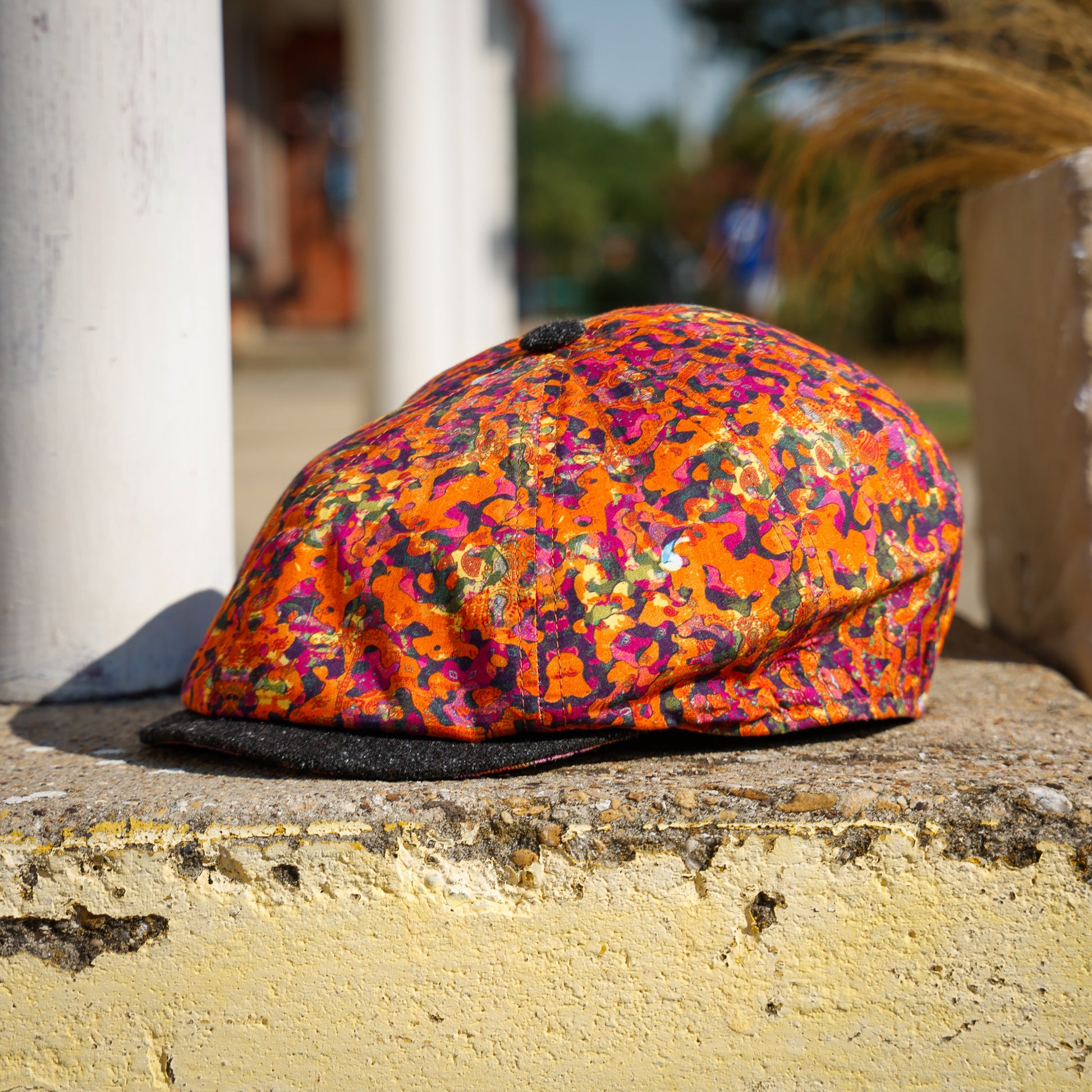 A flat cap with a vibrant orange, pink, and purple camouflage pattern, resting on a weathered stone ledge outdoors.