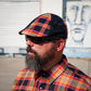 A man with a full beard wearing a black distressed flat cap with a vibrant orange, yellow, and red plaid pattern, along with dark sunglasses and a matching plaid button-down shirt, photographed outdoors against a white shuttered wall and a mural.