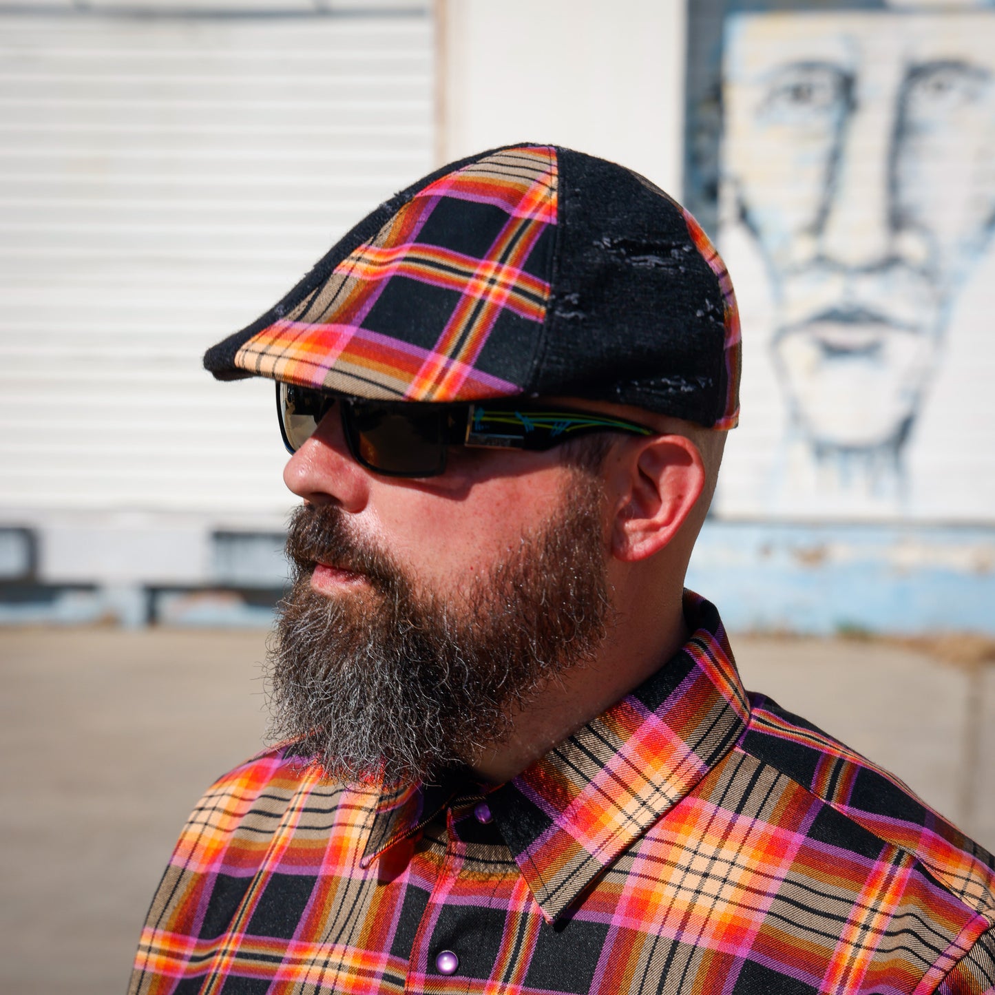 A man with a full beard wearing a black distressed flat cap with a vibrant orange, yellow, and red plaid pattern, along with dark sunglasses and a matching plaid button-down shirt, photographed outdoors against a white shuttered wall and a mural.