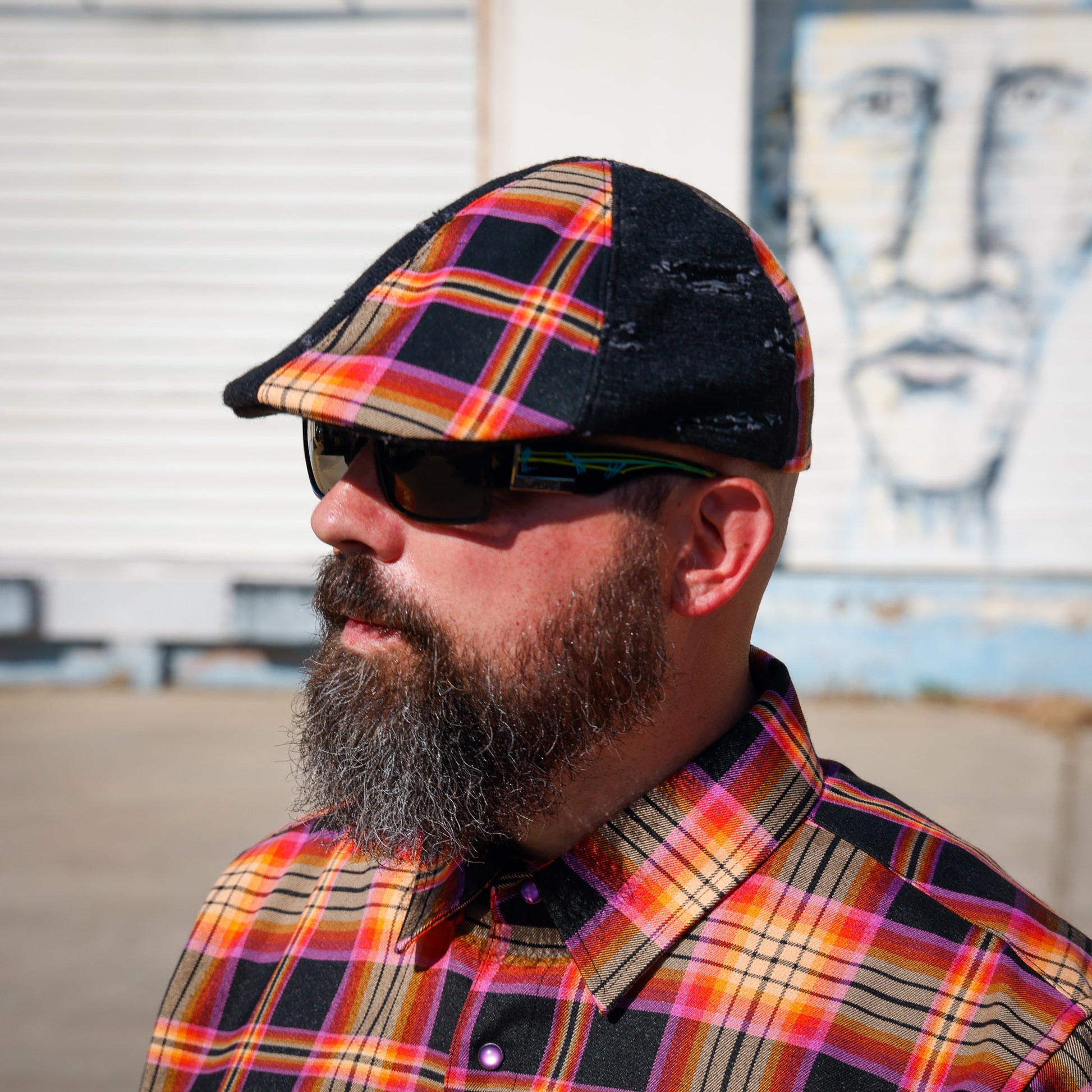 A man with a full beard wearing a black distressed flat cap with a vibrant orange, yellow, and red plaid pattern, along with dark sunglasses and a matching plaid button-down shirt, photographed outdoors against a white shuttered wall and a mural.