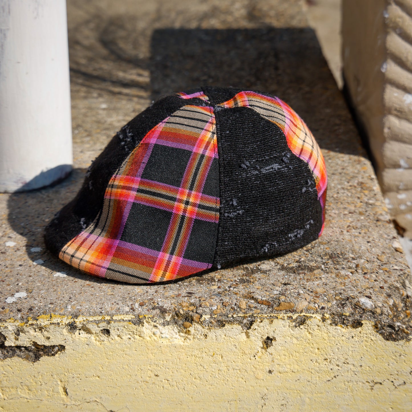 A flat-brim cap resting on a concrete ledge outdoors. The front panels feature a vibrant multicolor plaid pattern with orange, pink, yellow, and black stripes, while the side panels are black mesh.