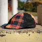 A baseball cap with a black distressed front panel and a colorful plaid side panel, resting on a weathered concrete ledge outdoors.