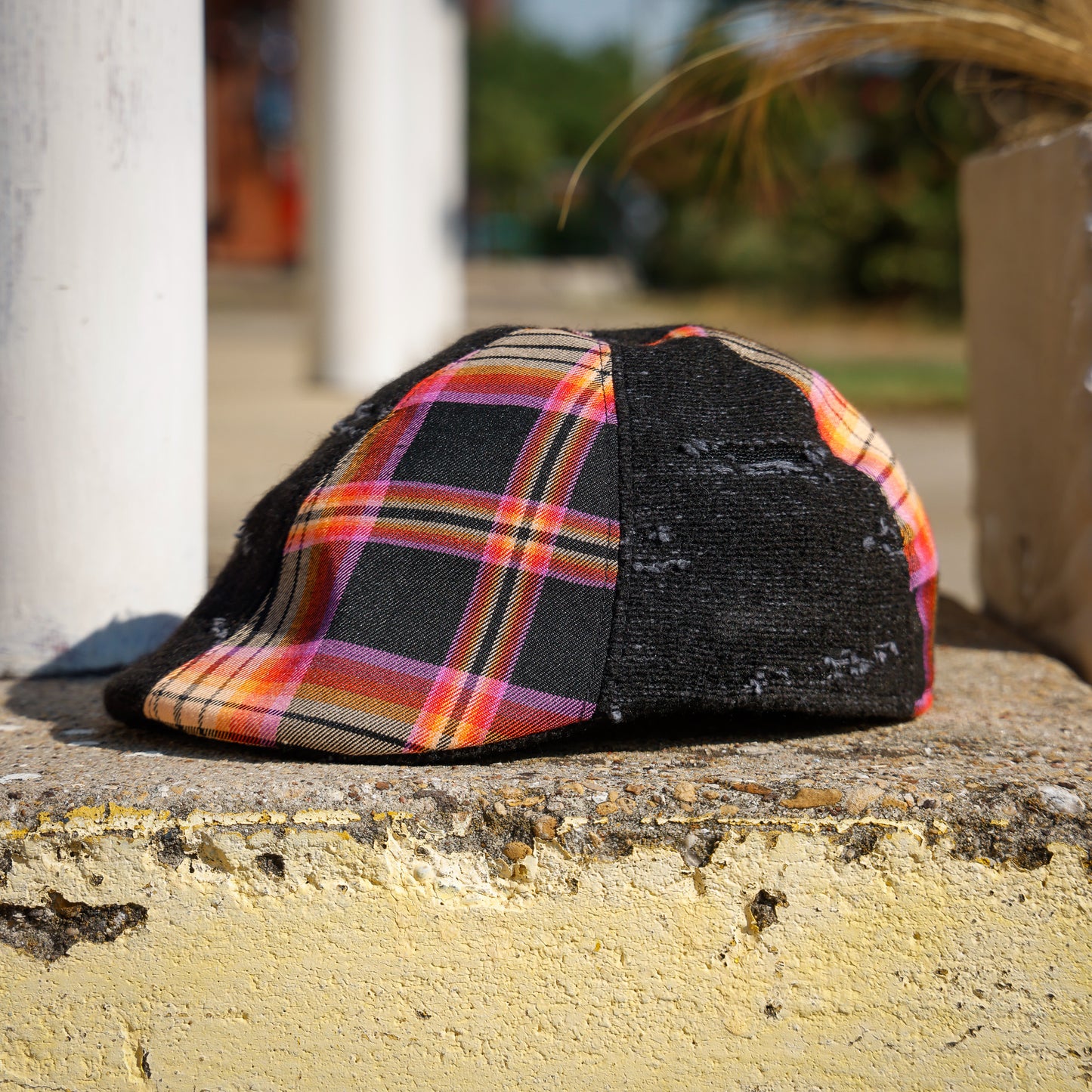 A baseball cap with a black distressed front panel and a colorful plaid side panel, resting on a weathered concrete ledge outdoors.