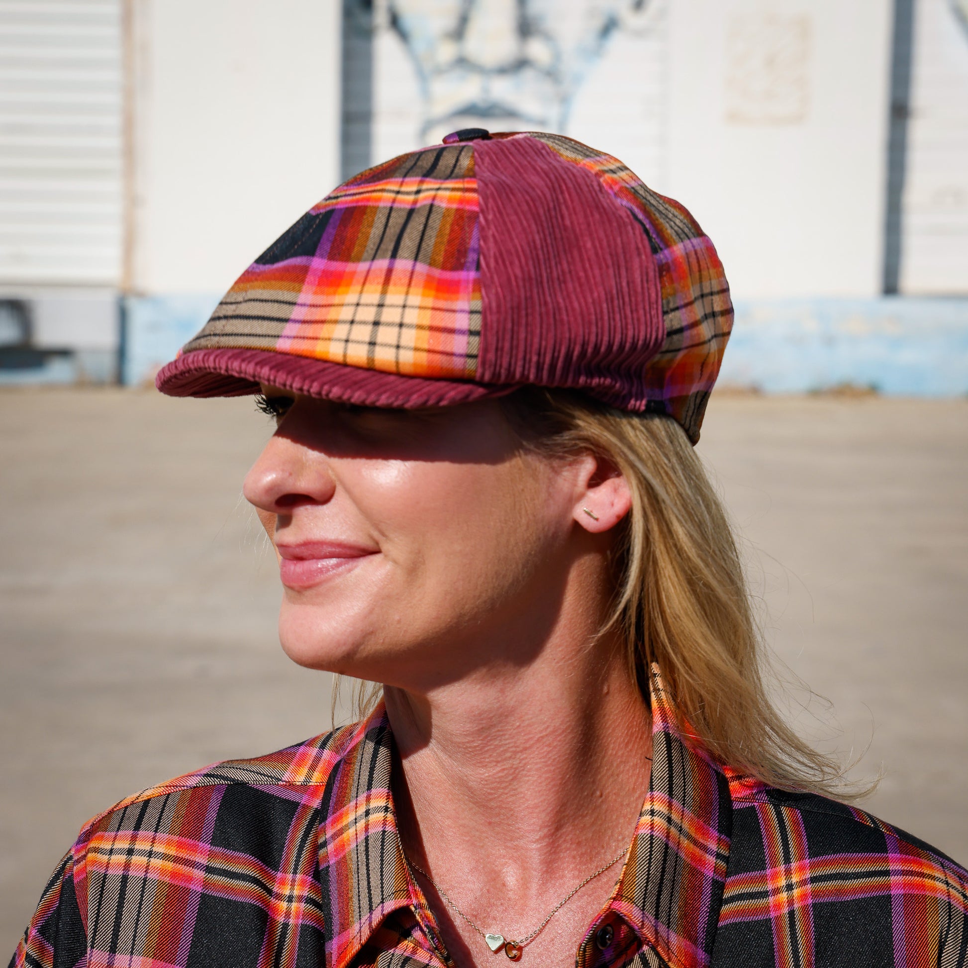 A woman wearing a plaid flat cap and matching plaid shirt, photographed outdoors in bright sunlight with a blurred building in the background.