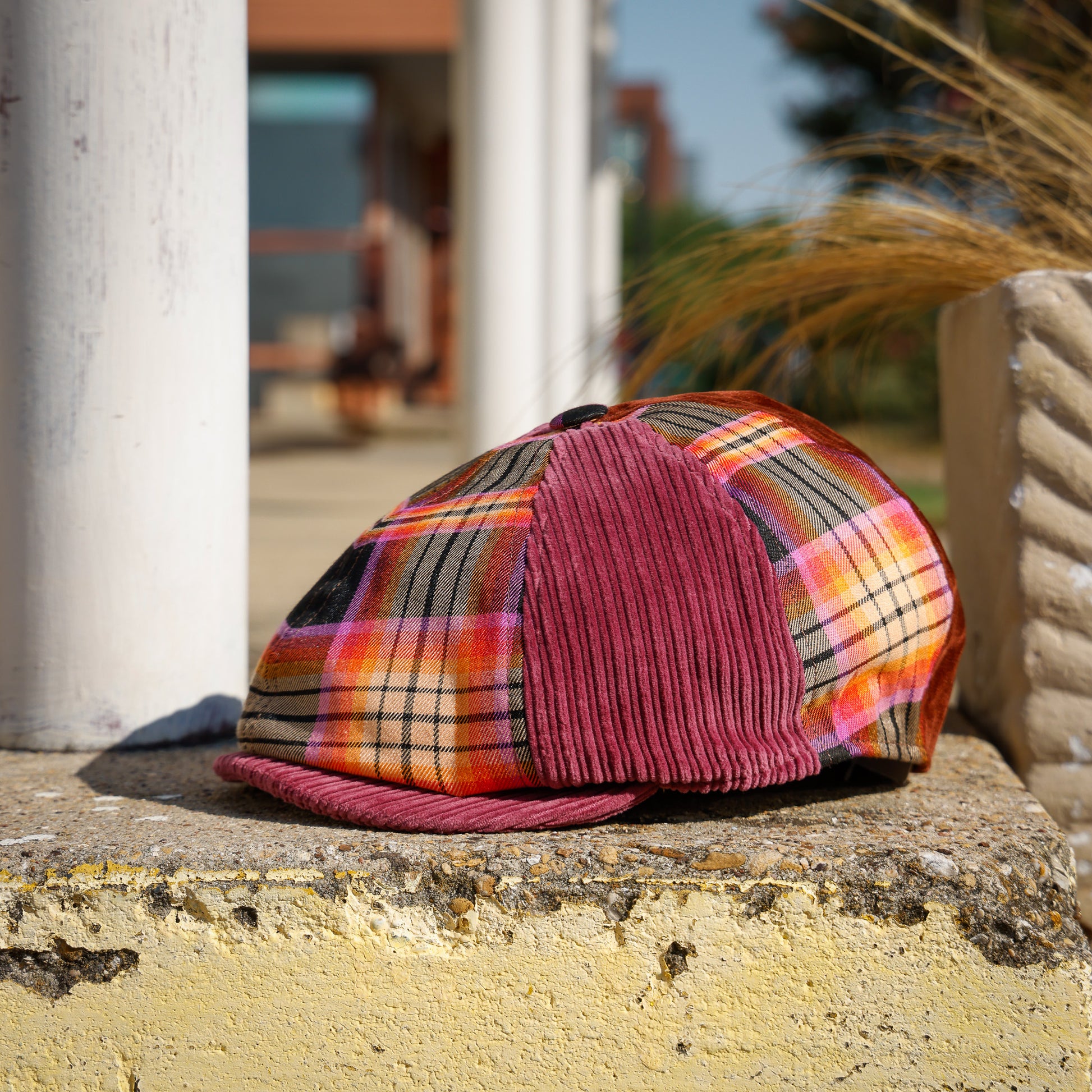 A multicolor plaid flat cap with a burgundy corduroy front panel and matching brim, resting on a weathered yellow stone ledge outdoors.