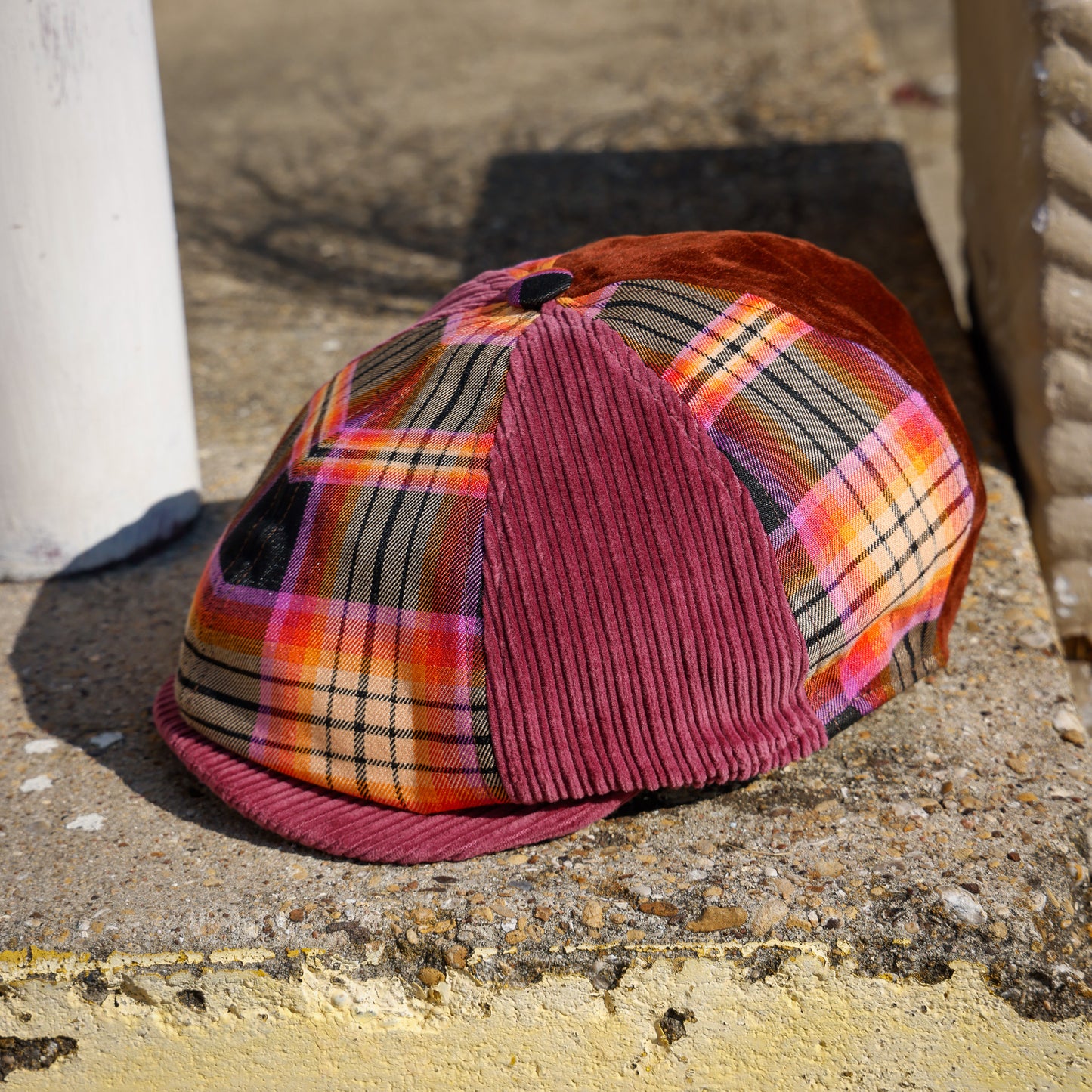 A vintage-style flat cap with a multicolor orange, pink, and black plaid front panel, a burgundy corduroy front visor, and a matching burgundy back, resting on a concrete ledge outdoors.