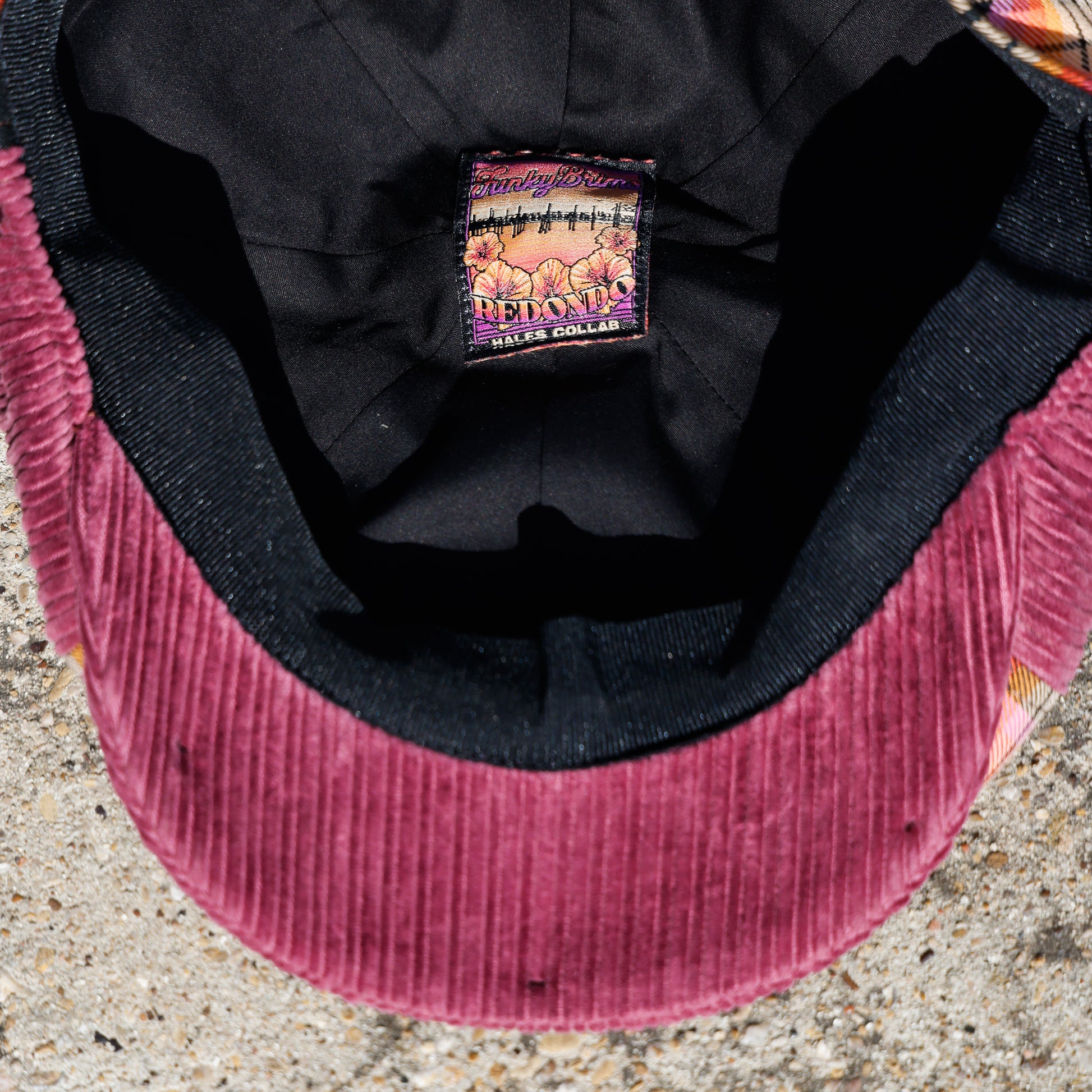 Close-up of the inside of a newsboy cap showing a burgundy corduroy brim, black fabric lining, and a colorful embroidered brand patch.