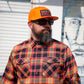 A man with a full beard and sunglasses wearing a bright orange baseball cap with 'HIVY' on the front, paired with a vibrant orange, yellow, and black plaid button-down shirt, standing in front of a white shutter and a blue-toned mural.