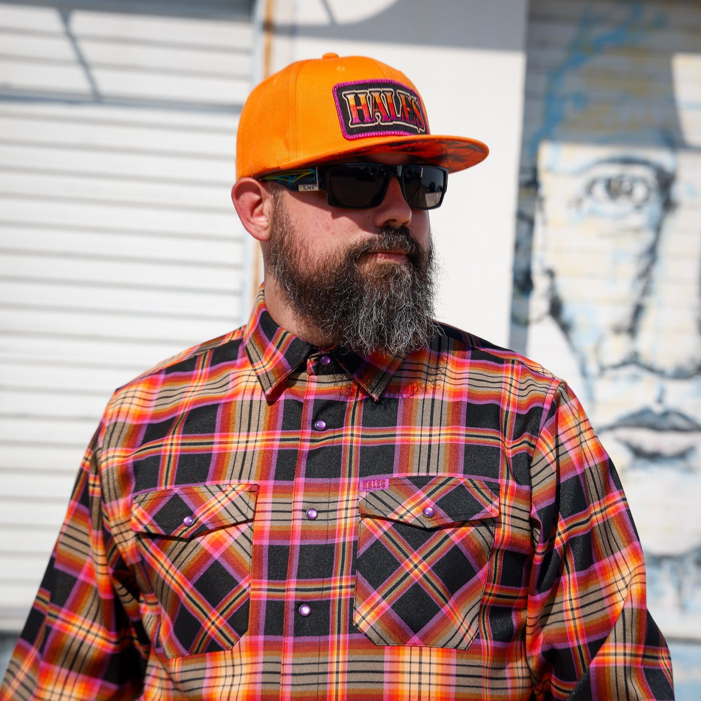 A man with a full beard and sunglasses wearing a bright orange baseball cap with 'HIVY' on the front, paired with a vibrant orange, yellow, and black plaid button-down shirt, standing in front of a white shutter and a blue-toned mural.