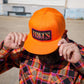 A man wearing a bright orange baseball cap with a 'HALES' patch on the front, holding the brim down over his face. He is wearing a colorful plaid flannel shirt, photographed outdoors against a blurred background.
