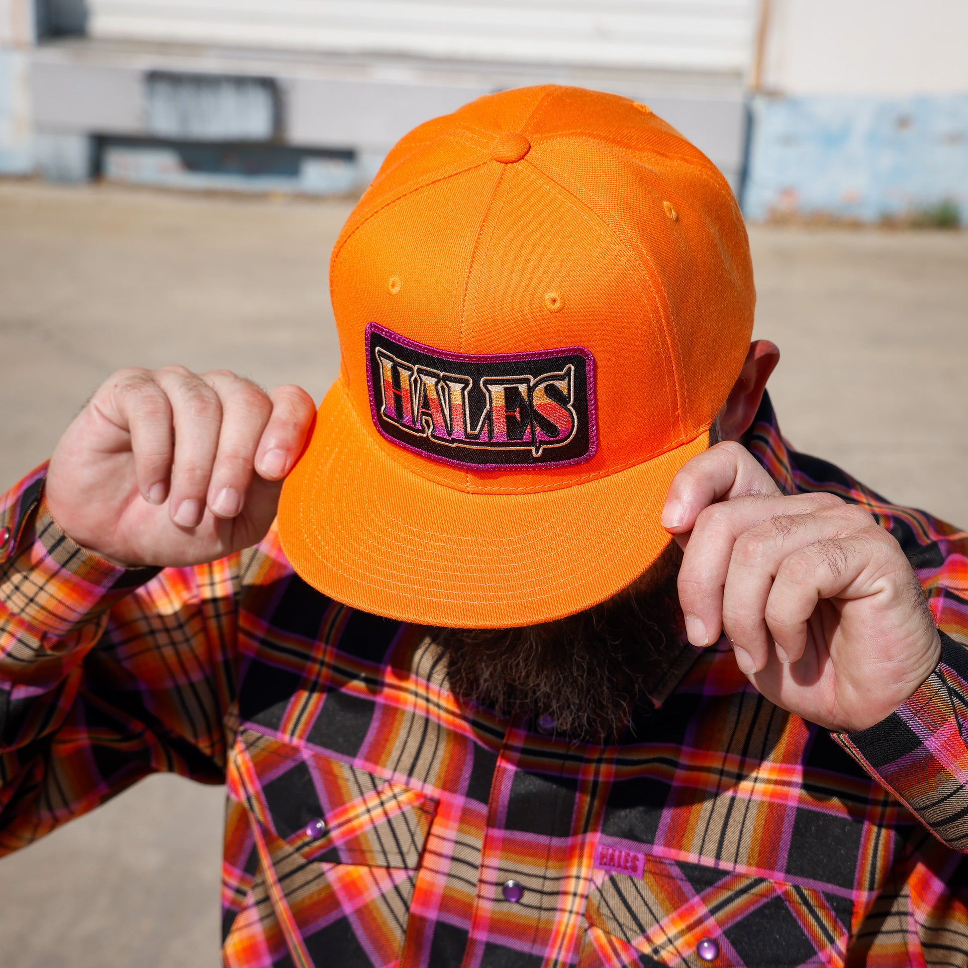 A man wearing a bright orange baseball cap with a 'HALES' patch on the front, holding the brim down over his face. He is wearing a colorful plaid flannel shirt, photographed outdoors against a blurred background.
