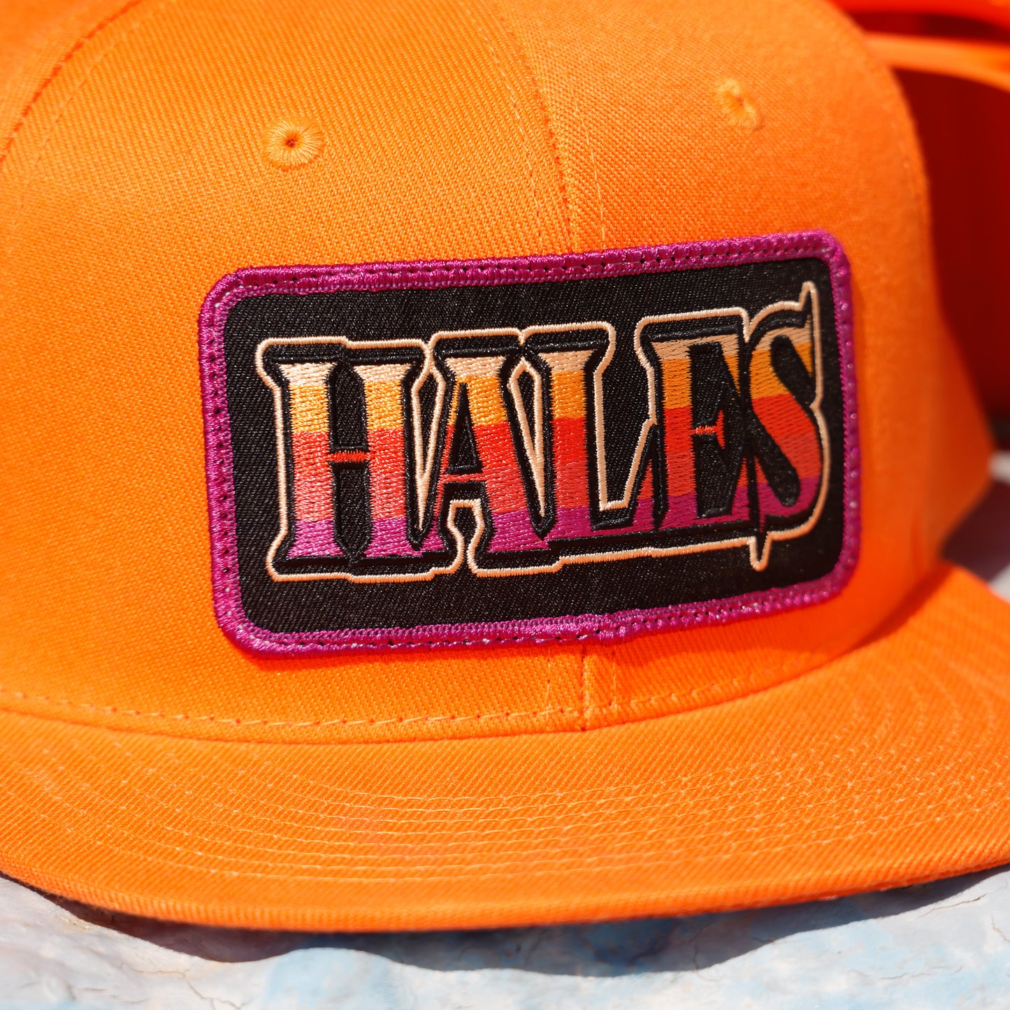 Close-up of a bright orange baseball cap featuring a rectangular embroidered patch with the word 'HALES' in stylized multicolor lettering on a black background with a purple border.