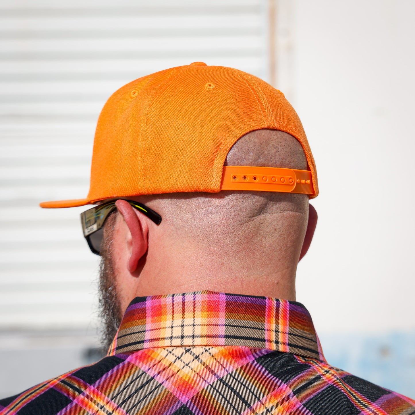 A man viewed from behind wearing a bright orange baseball cap and a colorful plaid shirt, with sunglasses on his face.