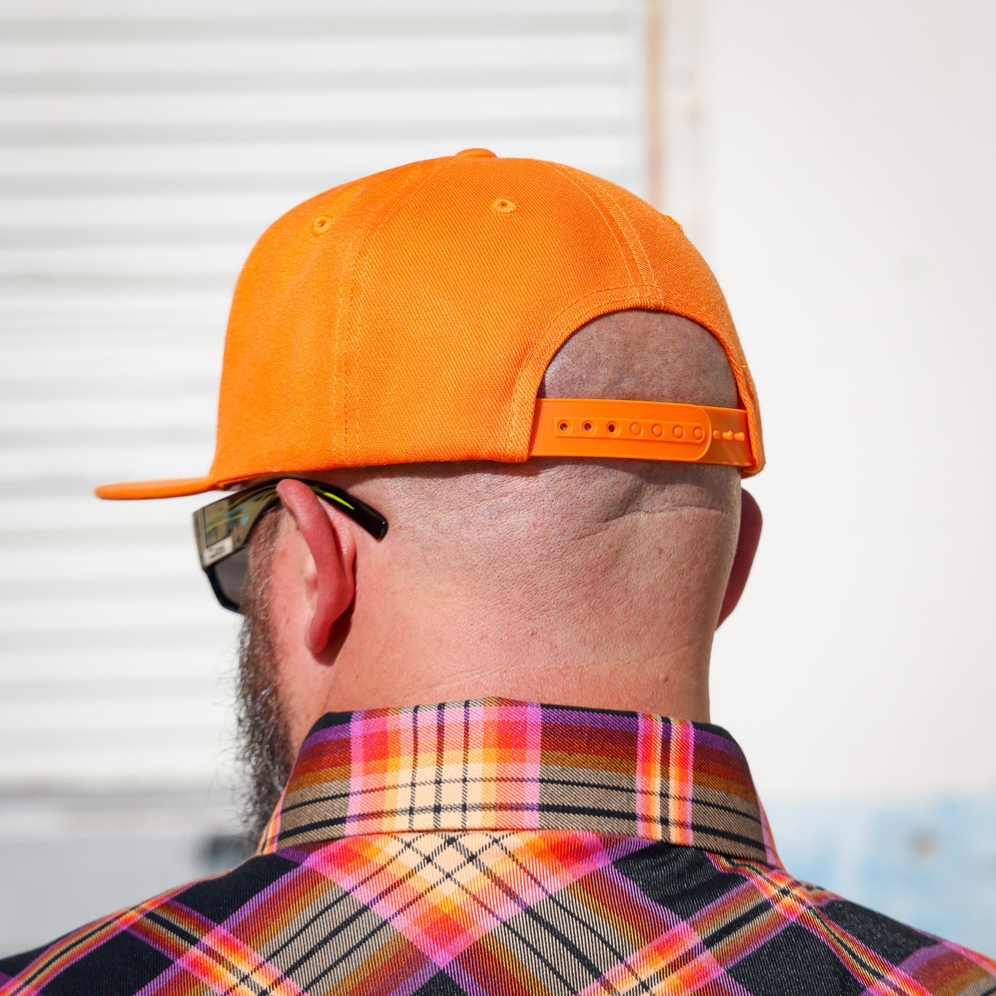 A man viewed from behind wearing a bright orange baseball cap and a colorful plaid shirt, with sunglasses on his face.