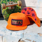 An orange snapback cap with a 'HALES' embroidered patch on the front, displayed next to another orange cap showing a purple and red floral patterned brim. A green skateboard is propped in the background against a weathered blue and white wall.