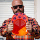 A man wearing a colorful plaid shirt and sunglasses holds up the inside of a bright orange and red baseball cap with a purple and orange floral patterned brim.