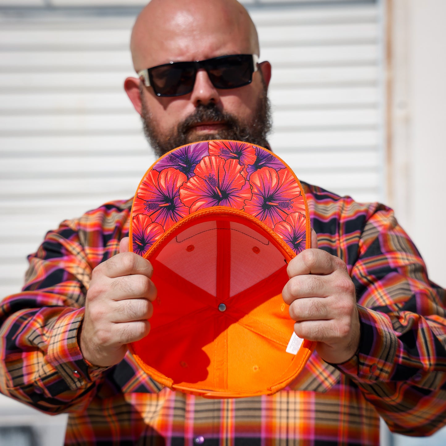 A man wearing a colorful plaid shirt and sunglasses holds up the inside of a bright orange and red baseball cap with a purple and orange floral patterned brim.