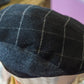 A close-up of a dark charcoal gray wool flat cap with a light beige windowpane plaid pattern, displayed on a mannequin head.