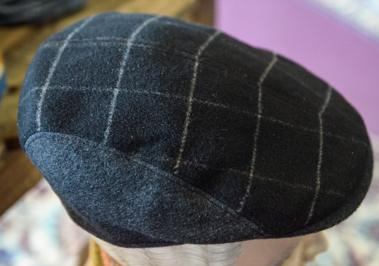 A close-up of a dark charcoal gray wool flat cap with a light beige windowpane plaid pattern, displayed on a mannequin head.