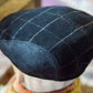 A close-up of a dark navy blue wool flat cap with a white windowpane plaid pattern, displayed on a mannequin head.