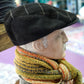 A mannequin bust wearing a black flat cap, a thick striped knit scarf in mustard, orange, cream, and gray, and a gray herringbone jacket, photographed indoors on a wooden surface.