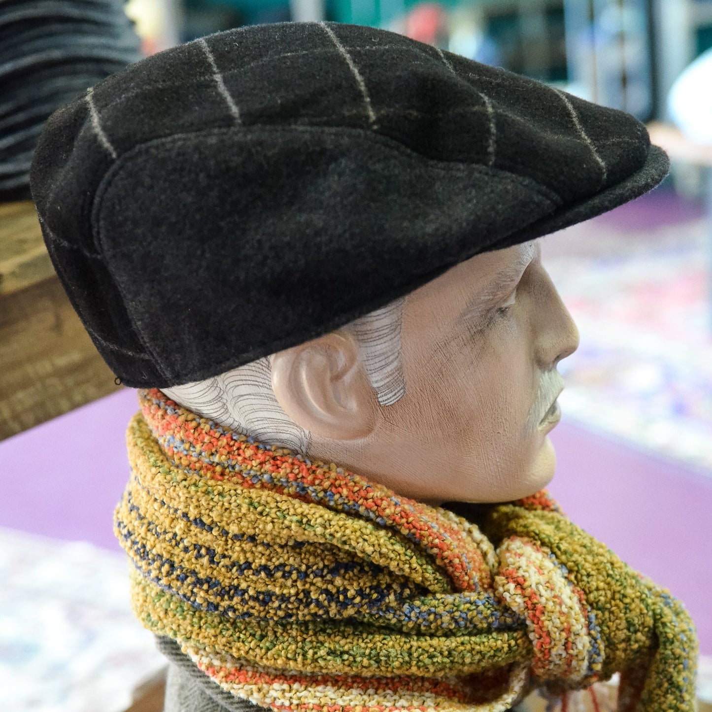 A mannequin bust wearing a black flat cap, a thick striped knit scarf in mustard, orange, cream, and gray, and a gray herringbone jacket, photographed indoors on a wooden surface.
