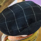 A close-up of a dark navy wool flat cap with a white windowpane plaid pattern, worn by a person with a striped knit scarf visible below.