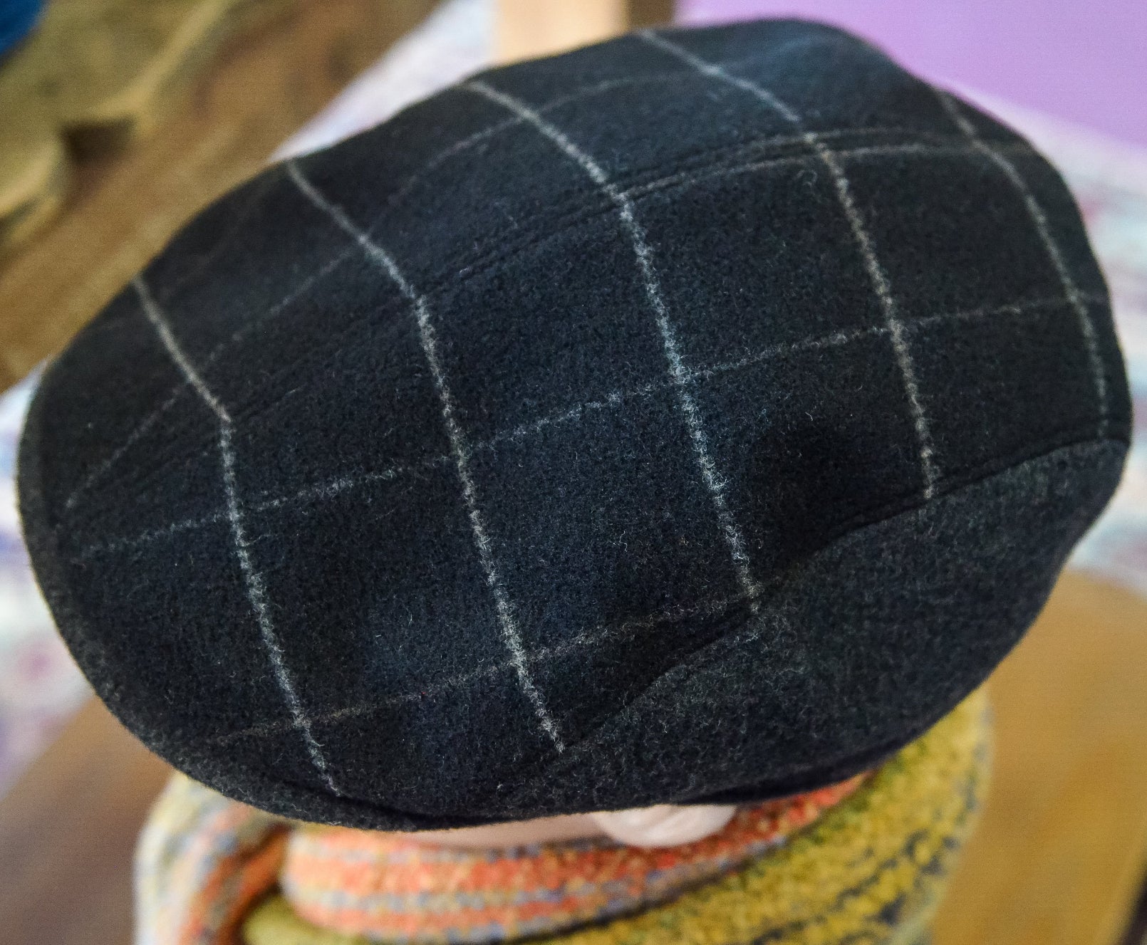 A close-up of a dark navy wool flat cap with a white windowpane plaid pattern, worn by a person with a striped knit scarf visible below.