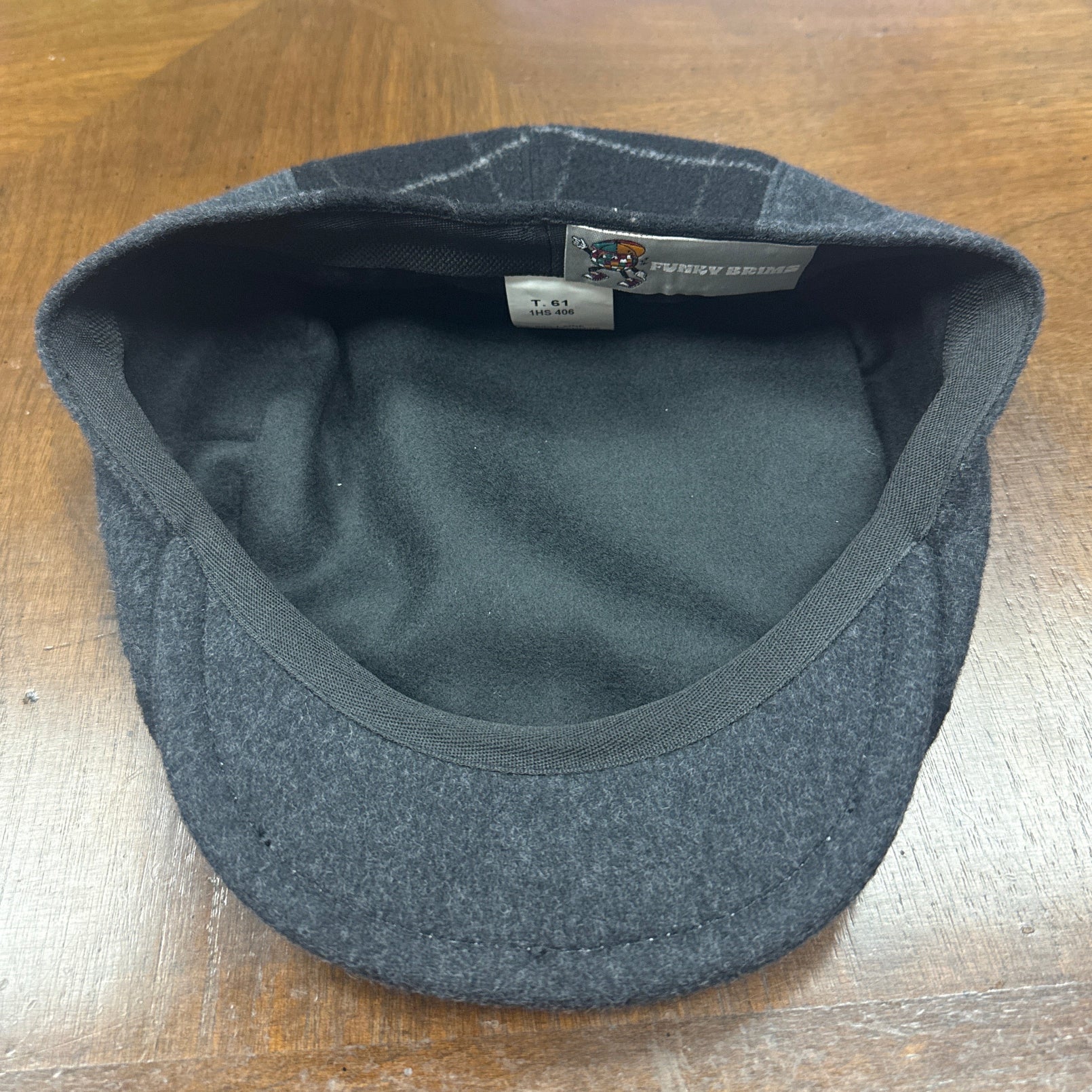 A dark gray wool flat cap with a black fleece interior, shown from the inside on a wooden desk. The brand label reads 'FunkyBrims.
