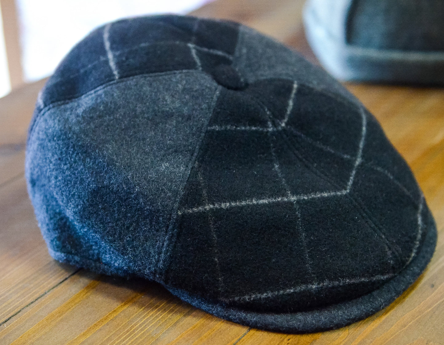 A dark charcoal gray wool flat cap with a subtle windowpane plaid pattern, resting on a wooden tabletop.