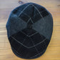 A dark gray and black checkered wool flat cap with a button top, photographed from above on a light brown wooden surface.