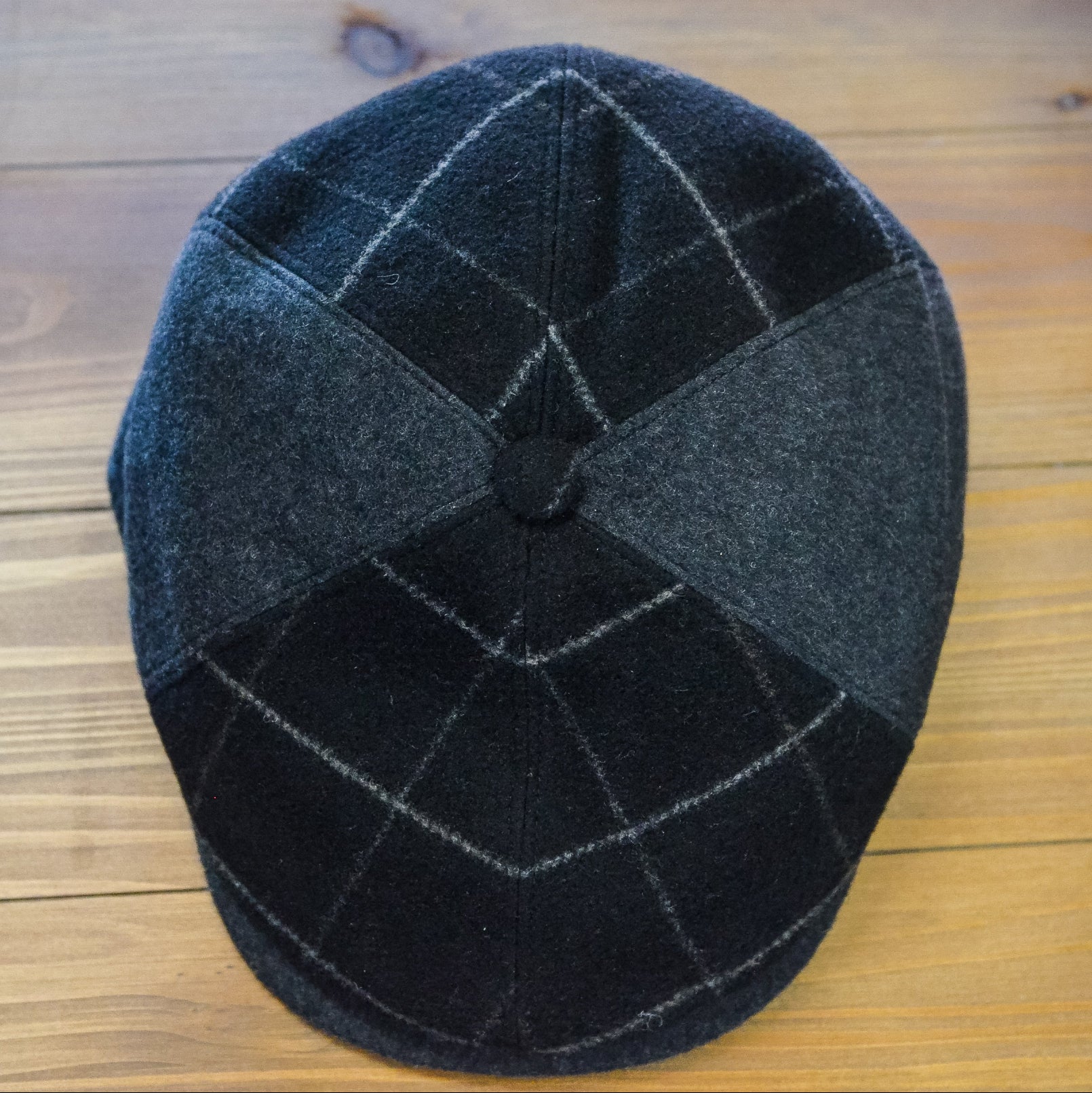 A dark gray and black checkered wool flat cap with a button top, photographed from above on a light brown wooden surface.