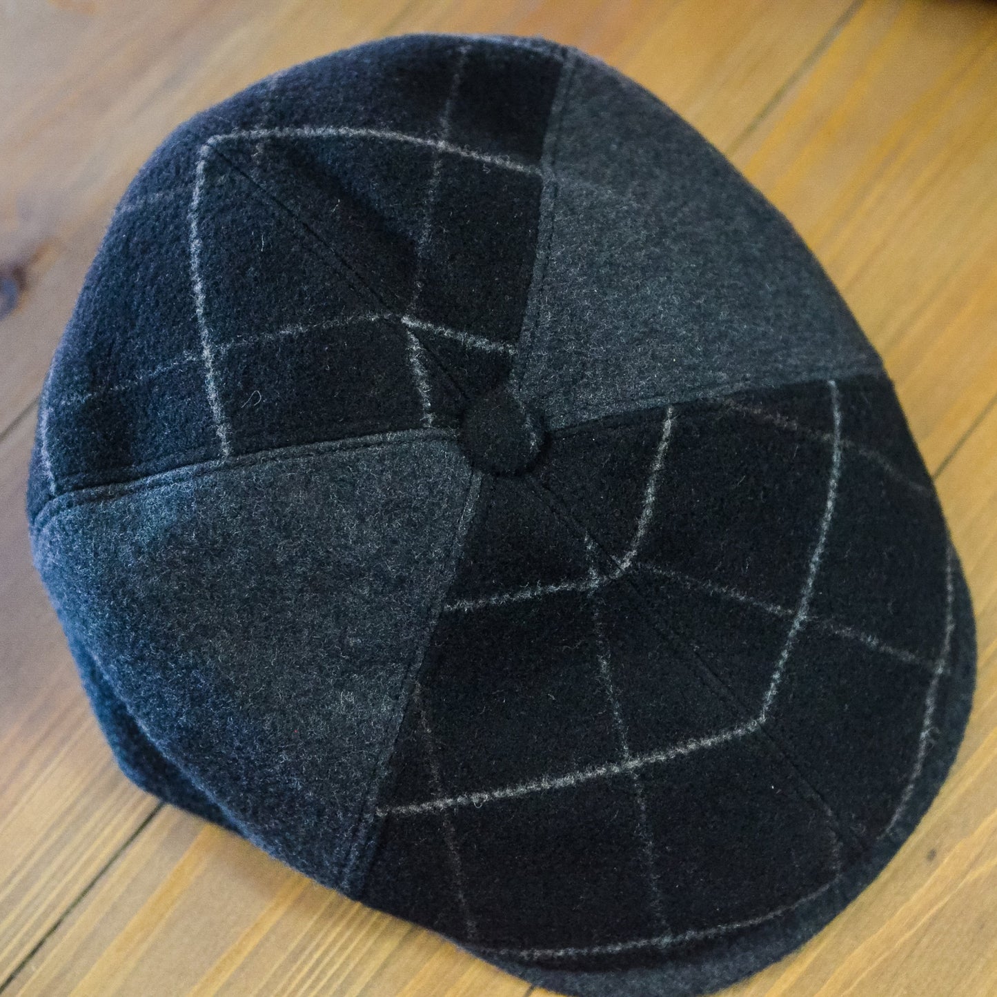 A dark gray and black patchwork flat cap with a subtle grid pattern, resting on a wooden surface.