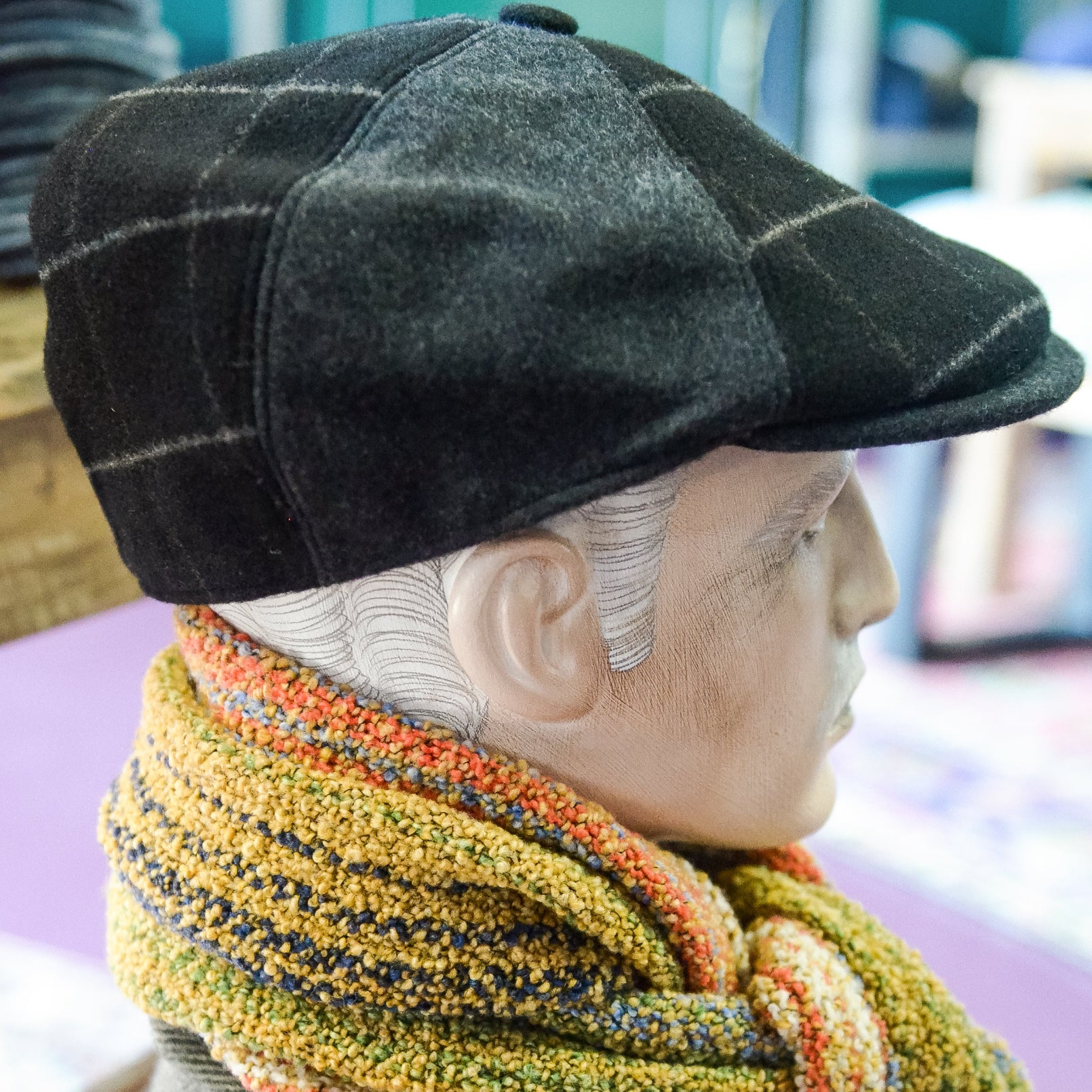 A close-up side profile of a mannequin or doll wearing a dark gray plaid flat cap, a thick knitted scarf in yellow, orange, and gray, and a brown herringbone jacket.