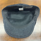 A dark gray wool flat cap with a black fleece lining, photographed from the inside view on a wooden table. A brand tag for is visible inside the crown.