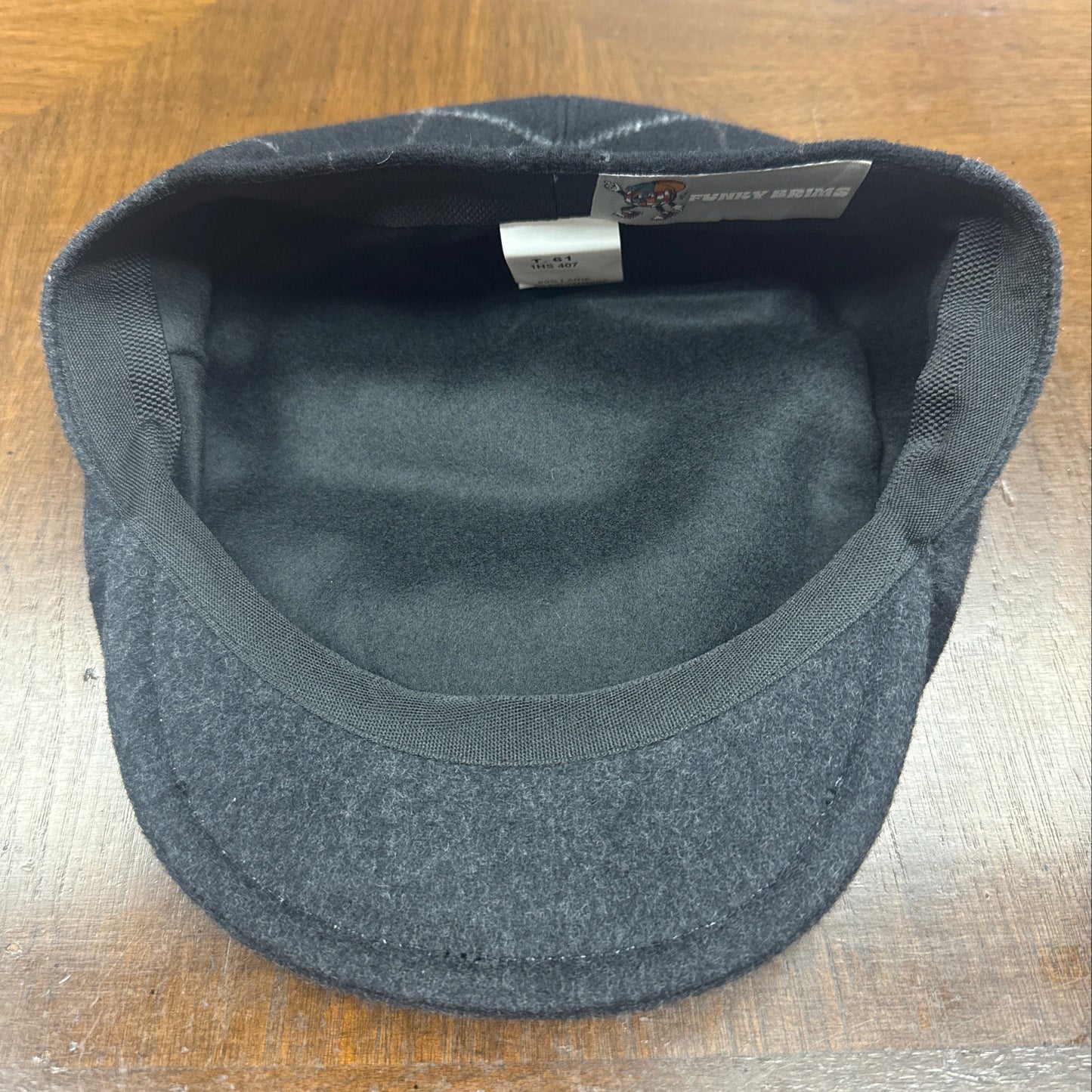 A dark gray wool flat cap with a black fleece lining, photographed from the inside view on a wooden table. A brand tag for is visible inside the crown.
