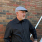 A man wearing a gray patterned flat cap and a black button-down shirt, standing in front of a brick wall.
