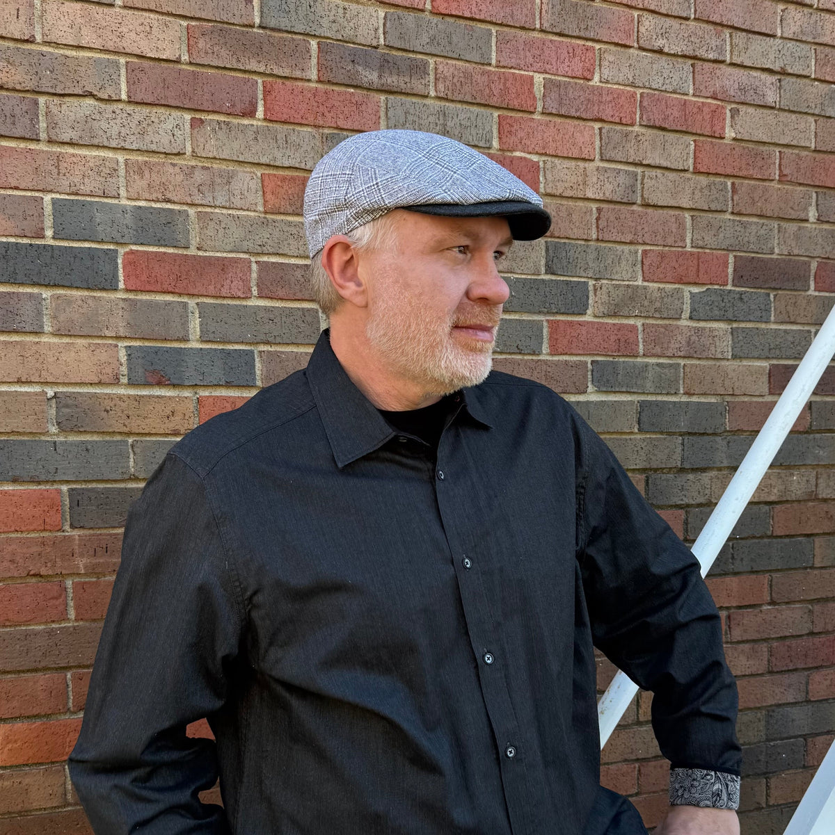 A man wearing a gray patterned flat cap and a black button-down shirt, standing in front of a brick wall.