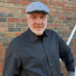A man wearing a gray herringbone flat cap and a dark charcoal gray button-down shirt, standing in front of a red and gray brick wall.
