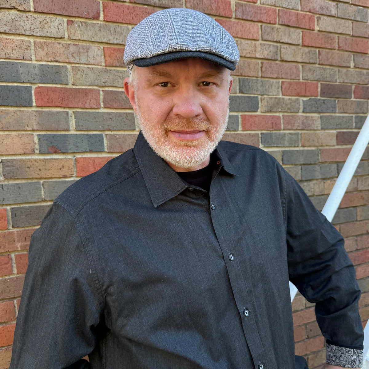 A man wearing a gray herringbone flat cap and a dark charcoal gray button-down shirt, standing in front of a red and gray brick wall.