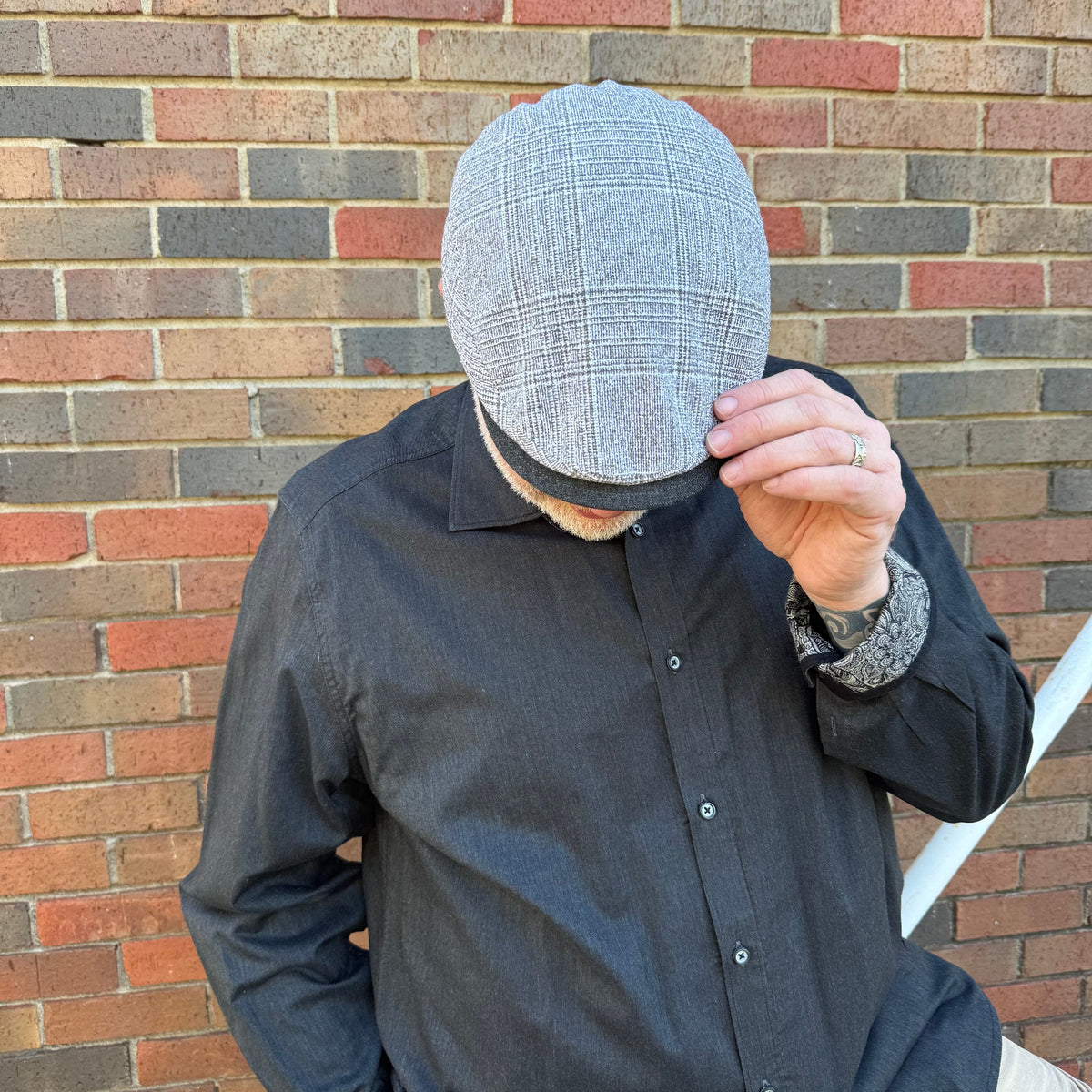 A man wearing a gray plaid flat cap and a dark charcoal button-down shirt stands in front of a red and gray brick wall, holding the brim of the cap with his hand.