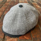 A gray and white plaid newsboy cap with a dark gray visor and button, resting on a brick surface.