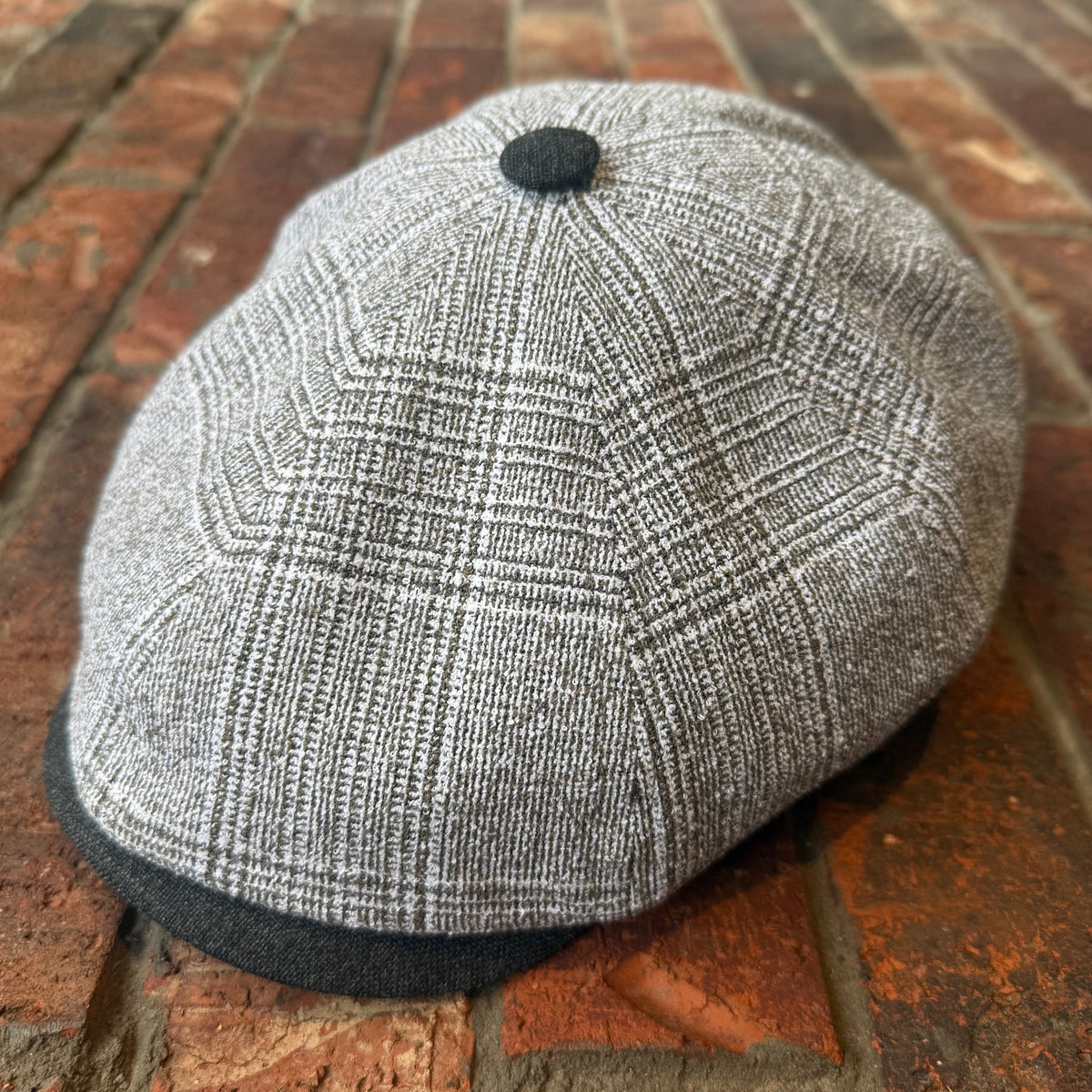 A gray and white plaid newsboy cap with a dark gray visor and button, resting on a brick surface.