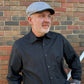 A man wearing a gray flat-brim cap and a dark charcoal button-down shirt, standing in front of a red and gray brick wall.