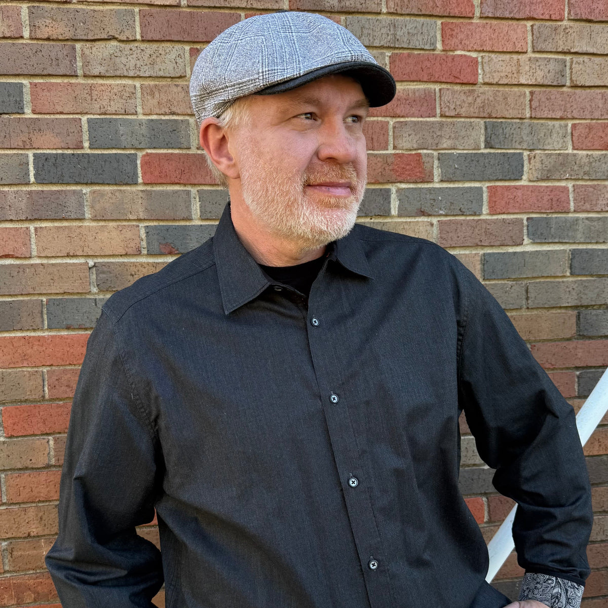 A man wearing a gray flat-brim cap and a dark charcoal button-down shirt, standing in front of a red and gray brick wall.