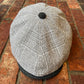 A gray and white plaid flat cap with a dark gray visor and top button, photographed from above on a red brick surface.