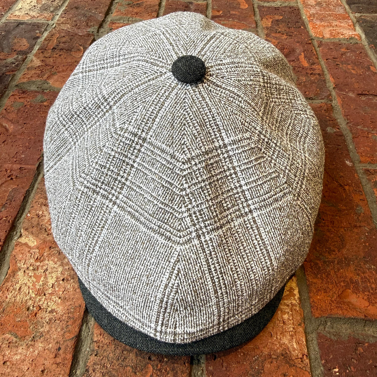 A gray and white plaid flat cap with a dark gray visor and top button, photographed from above on a red brick surface.