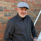 A man wearing a gray patterned flat cap and a dark charcoal button-down shirt, standing in front of a red and gray brick wall.