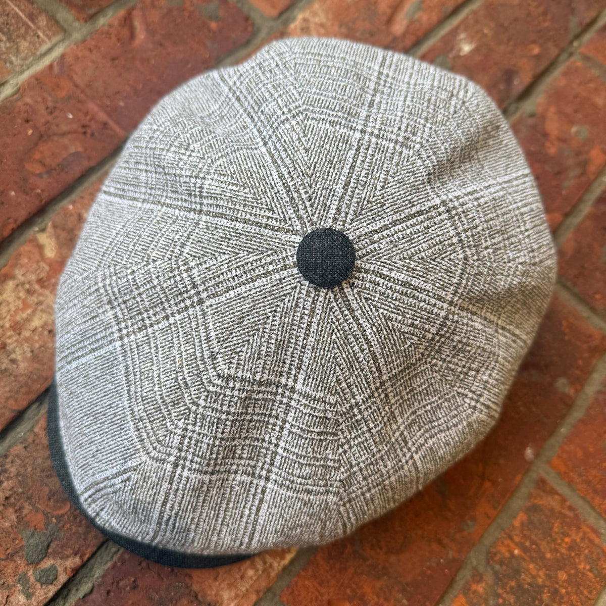 A gray and white plaid flat cap with a black button on top, resting on a red brick surface.